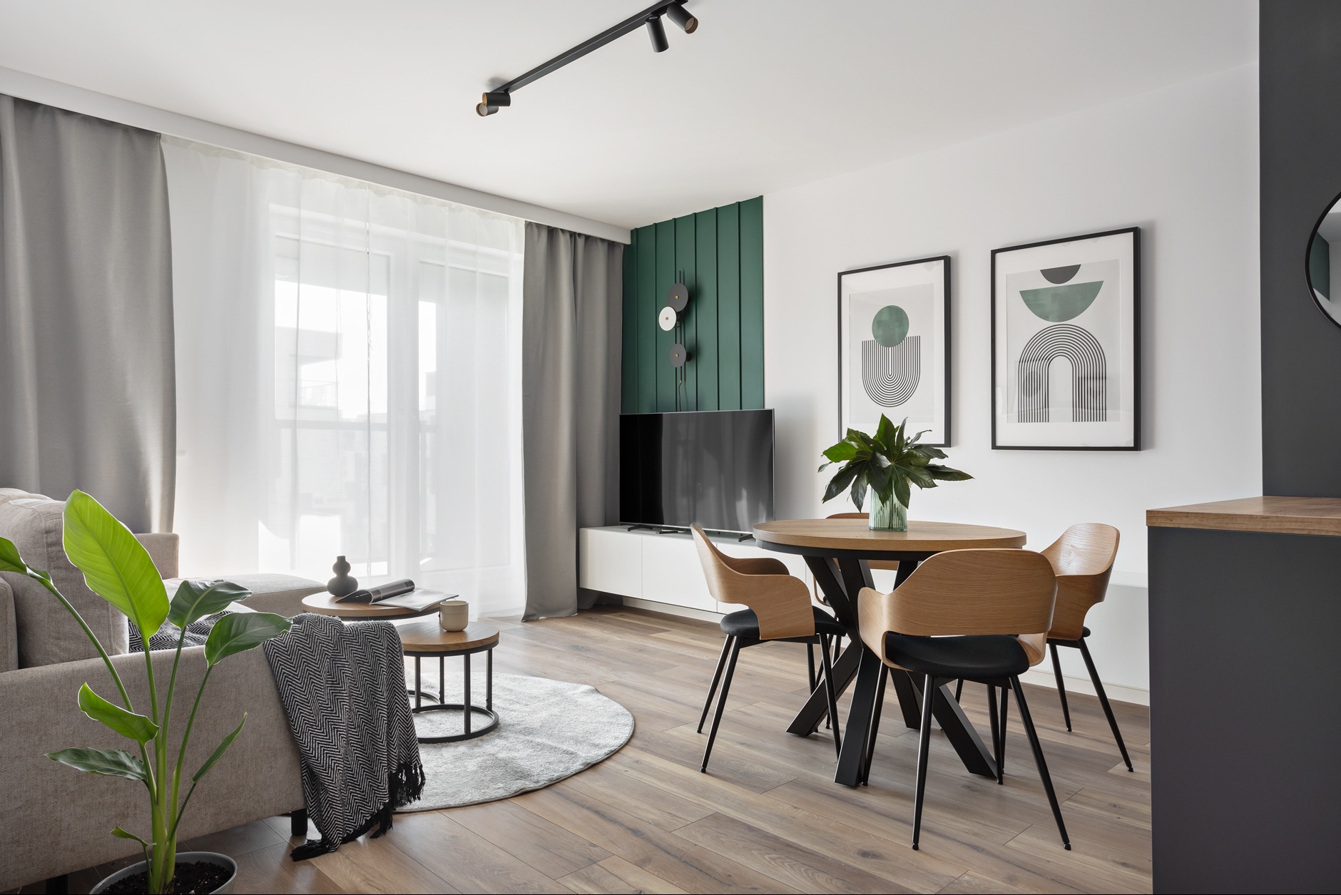 Jasne wnętrze salonu z drewnianą podłogą, okrągłym stołem i krzesłami. Na ścianie grafiki w czarnych ramkach i zielony akcent dekoracyjny. Widoczne rośliny doniczkowe.