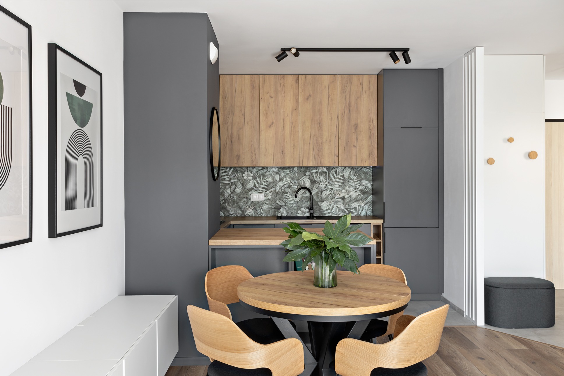 Stylowa aranżacja kuchni z drewnianymi szafkami, szarymi ścianami i okrągłym stołem z krzesłami. Uwagę przyciągają grafiki na ścianie i tropikalny wzór nad blatem.