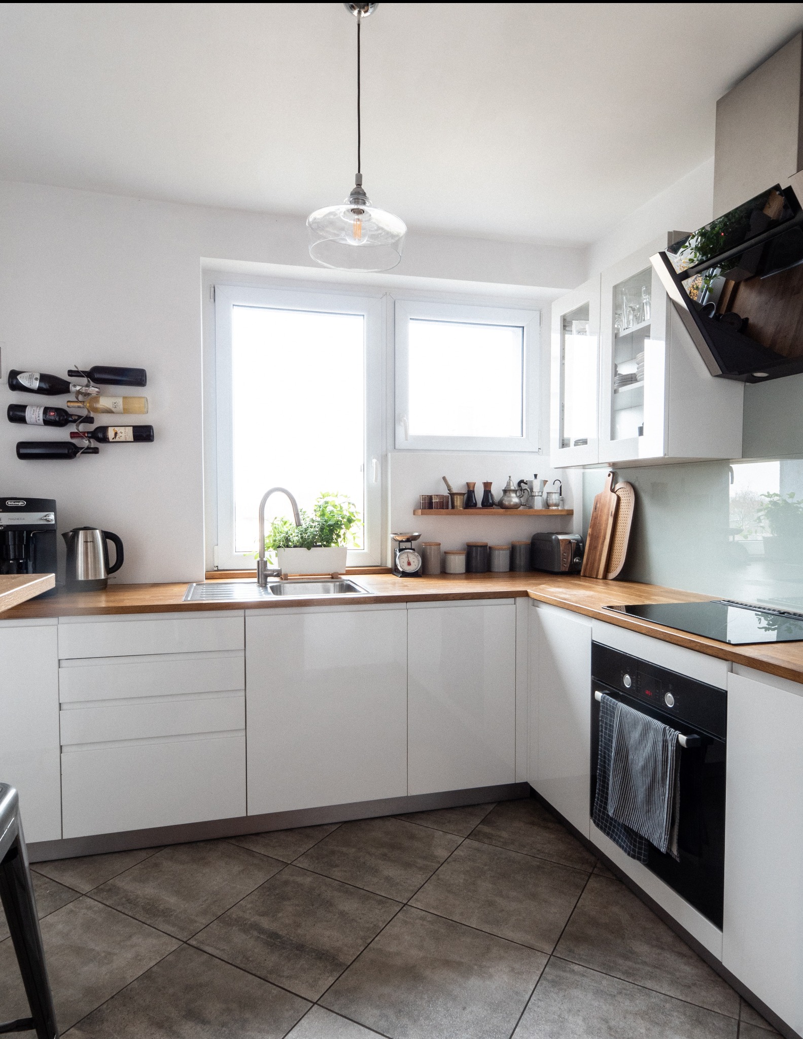 Jasna kuchnia w stylu skandynawskim z drewnianym blatem, białymi szafkami i szarymi płytkami podłogowymi. Na blacie ekspres do kawy, przyprawy i zioła w doniczce.