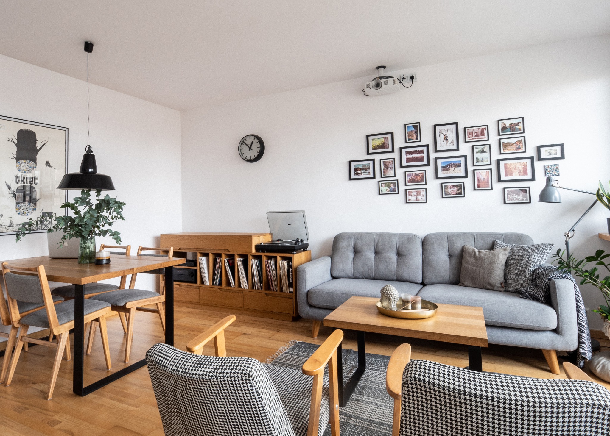 Jasne wnętrze salonu z drewnianym stołem, szarą sofą i fotelami w pepitkę. Na ścianie galeria zdjęć i projektor. Drewniana podłoga i regał z płytami.