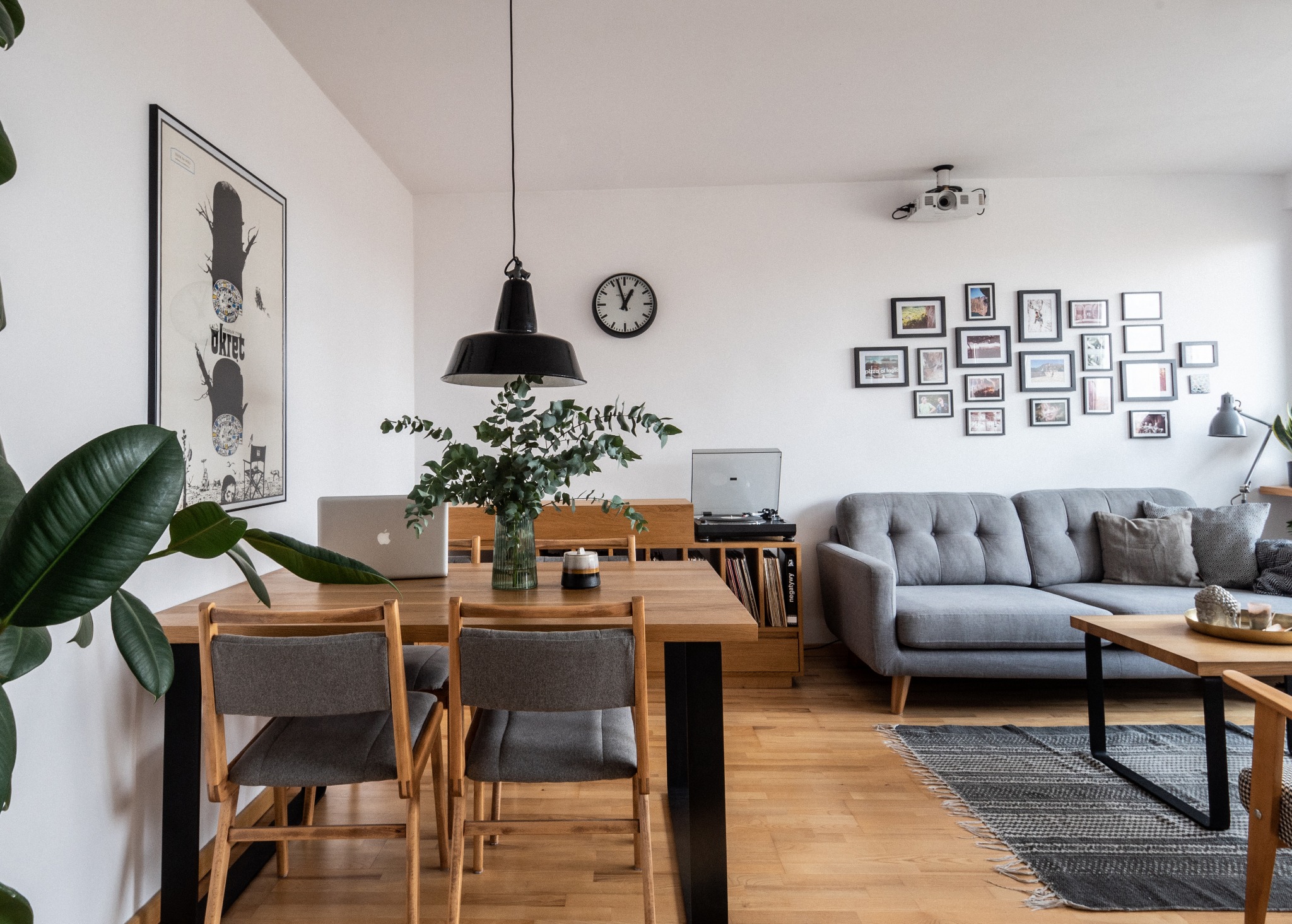 Jasne, przestronne wnętrze: drewniany stół z laptopem i wazonem, szara sofa z poduszkami, galeria zdjęć na ścianie, gramofon i dywan w stylu vintage.