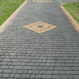 Długa ścieżka wyłożona szarą kostką brukową z dekoracyjnymi elementami w kształcie rombu i okręgu, obrzeża z kostki w odcieniach żółci i pomarańczy, trawnik po bokach.
