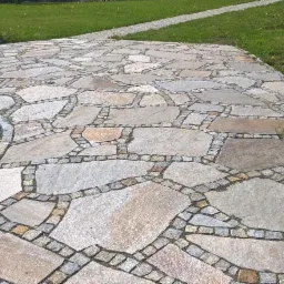 Nieregularny wz&oacute;r z jasnoszarego kamienia na ścieżce ogrodowej, obramowany drobną kostką, z trawnikiem w tle.