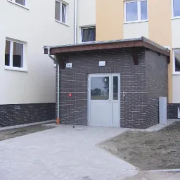 Zewnętrzny widok przyłącza budynku z instalacją odgromową, widoczne elementy uziemiające i przewód odprowadzający na elewacji, obok skrzynka elektryczna.