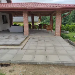 Układanie szarej kostki brukowej na tarasie z ceglanymi filarami, widoczne niedokończone prace ziemne i przygotowanie podłoża.