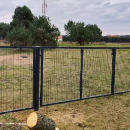 Zestaw ciemnoszarej bramy wjazdowej i przęseł z siatki zgrzewanej na tle łąki i lasu iglastego, widoczne ścięte gałęzie i pniak.