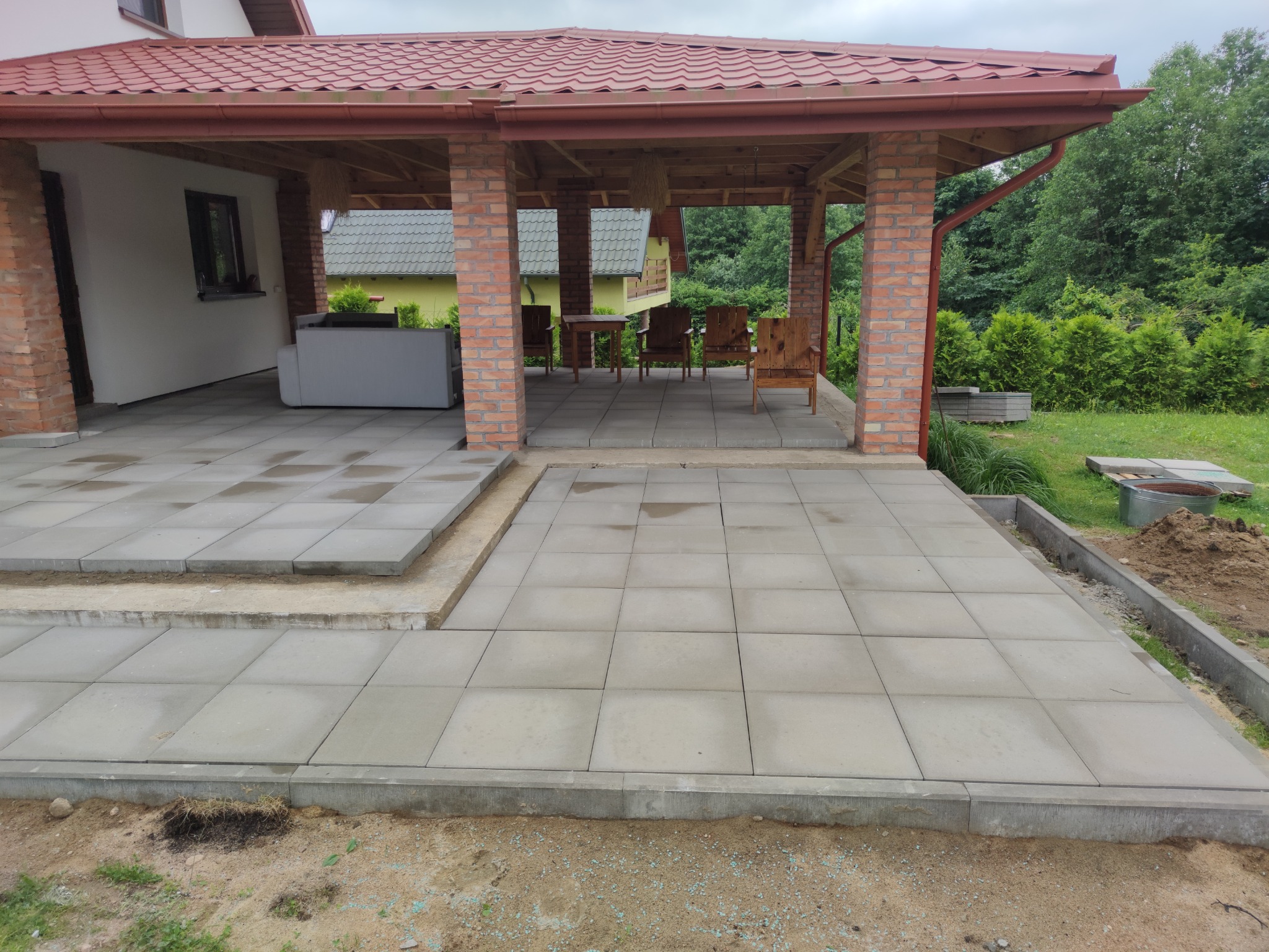 Układanie szarej kostki brukowej na tarasie z ceglanymi filarami, widoczne niedokończone prace ziemne i przygotowanie podłoża.