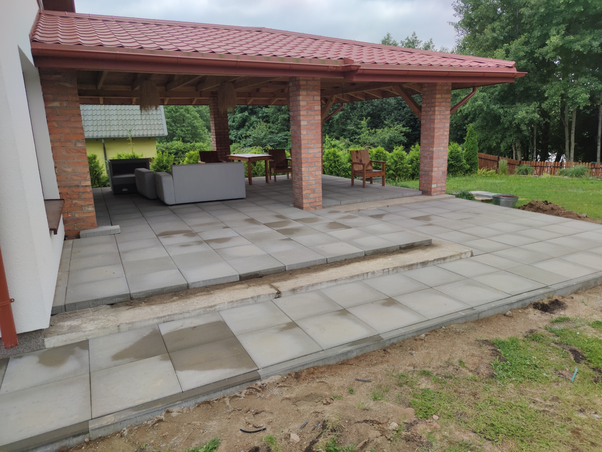 Układanie szarych płyt chodnikowych na tarasie z ceglanymi filarami pod zadaszeniem, widoczne nierówności terenu i prace wykończeniowe.