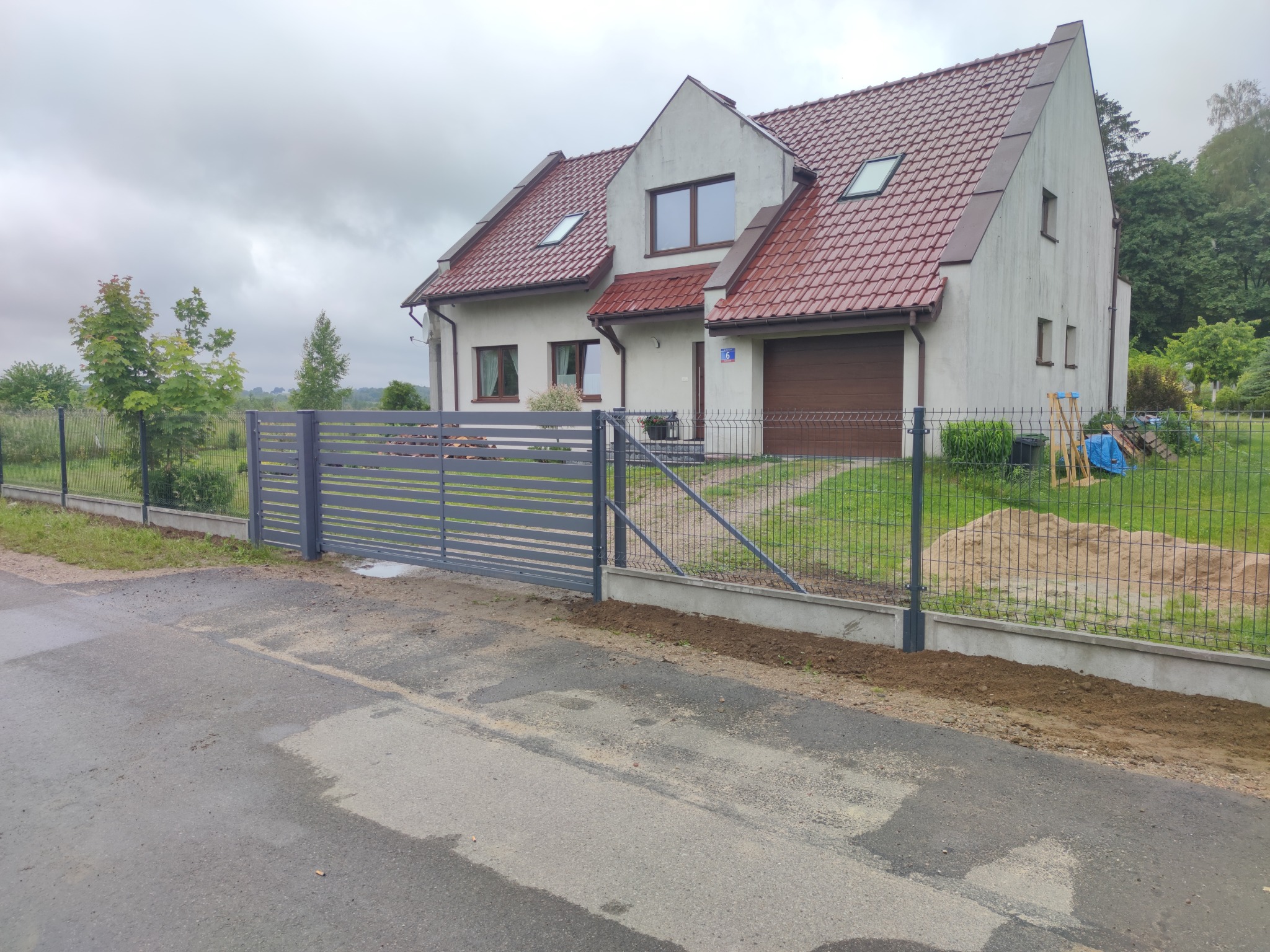 Nowoczesna posesja z szarą przesuwną bramą wjazdową i ogrodzeniem z siatki, widok na dom z czerwoną dachówką w pochmurny dzień.