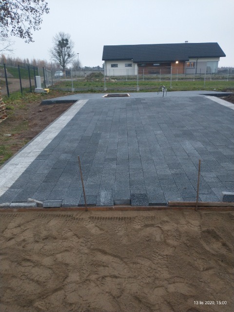 Układanie szarej kostki brukowej na podsypce piaskowej, widoczne drewniane obramowanie i wystające pręty, w tle dom jednorodzinny i drzewa.