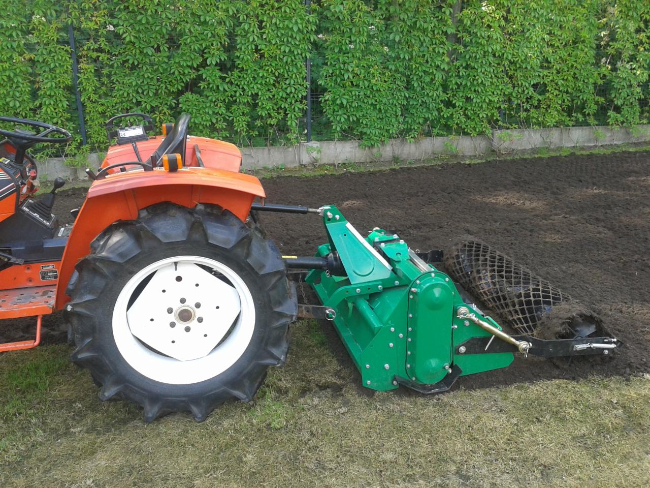 Pomarańczowy ciągnik z zielonym agregatem uprawowym separacyjnym przygotowuje glebę na trawniku, widoczna świeżo uprawiona ziemia i siatka separacyjna.