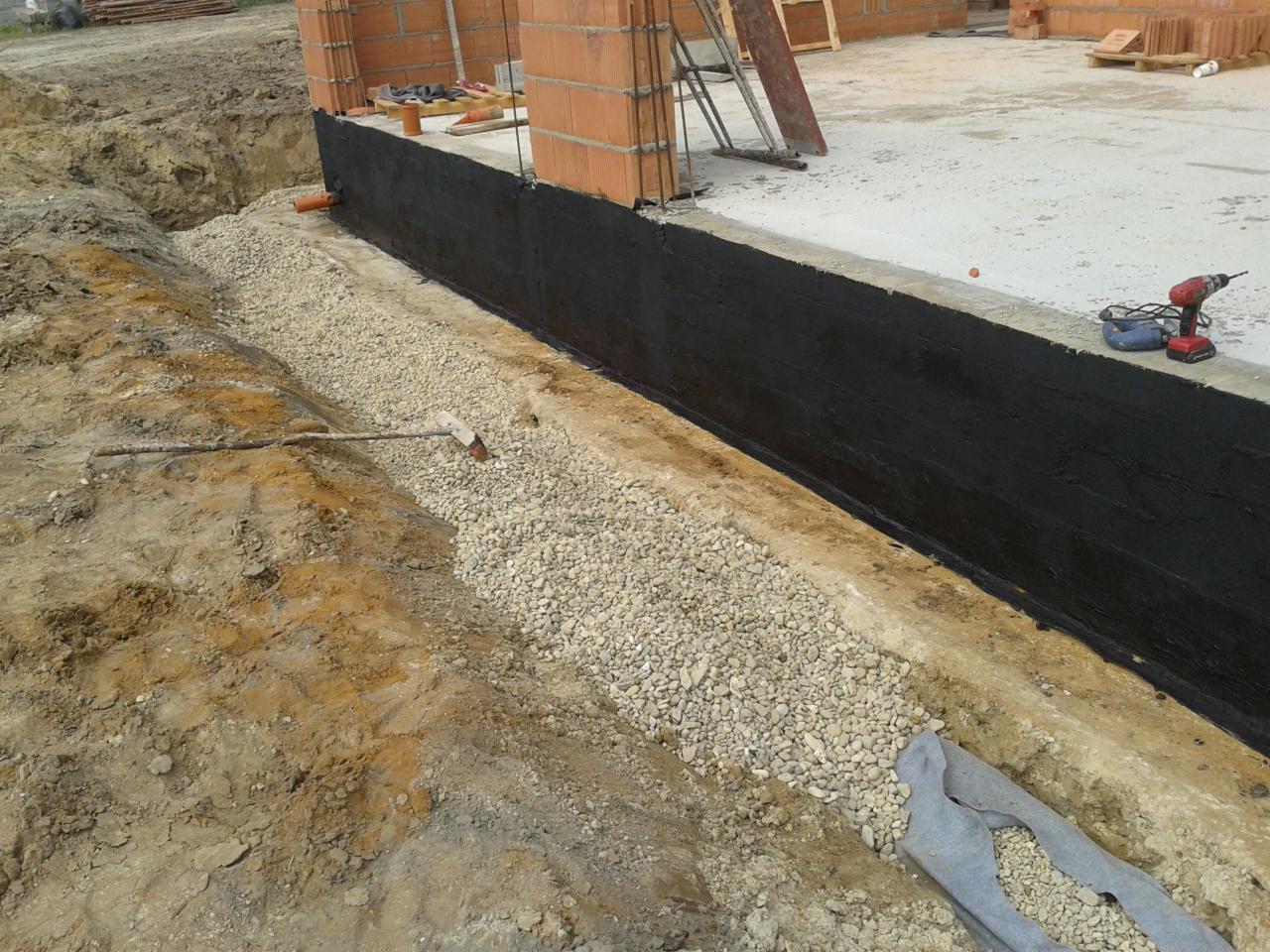 Fundament budynku w trakcie budowy, zabezpieczony czarną izolacją przeciwwilgociową, z warstwą drenażową z kamieni i częściowo przykryty geowłókniną. W tle ceglany mur i narzędzia.