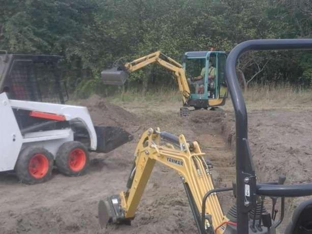 Dwa żółte minikoparki Yanmar i biała ładowarka Bobcat pracują przy wykopie w terenie trawiastym, z widocznym fragmentem kabiny jednej z koparek.