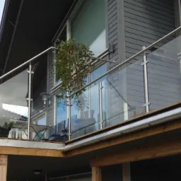 Szklana balustrada z nierdzewnymi słupkami na balkonie domu z szarą elewacją, widok z dołu.
