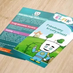 Dwie ulotki reklamowe Robo Elfik leżące na jasnym drewnianym biurku, z bliska widoczne informacje o zajęciach z robotyki, programowania Scratch i szach&oacute;w dla dzieci w Raciborzu.