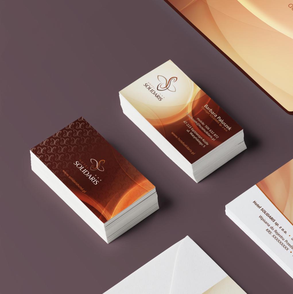 Stos wizytówek z logo i danymi kontaktowymi Hotelu Solidaris, ułożone na szarym tle obok koperty i materiałów reklamowych w ciepłej tonacji brązowo-pomarańczowej.