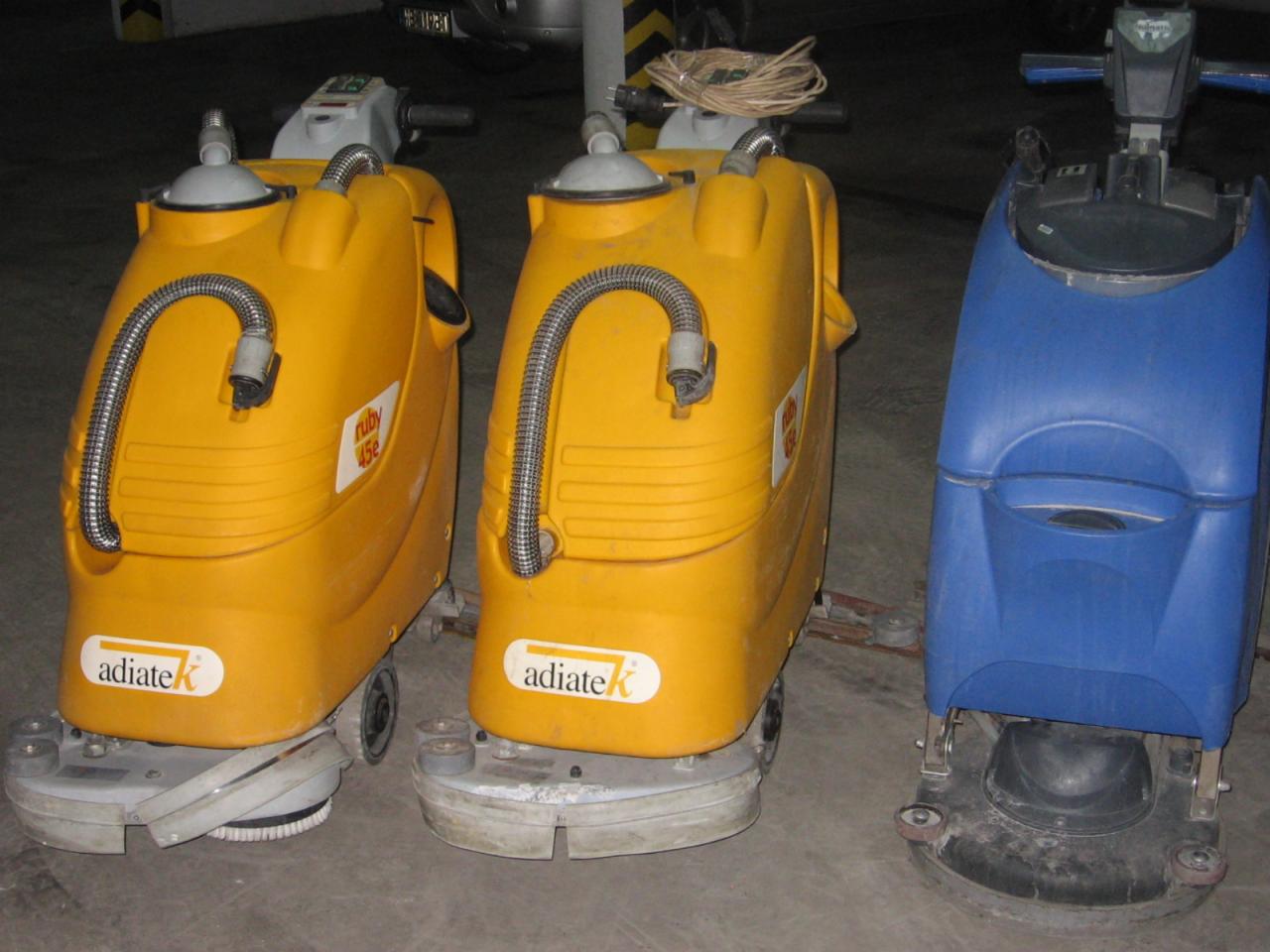 Trzy maszyny do czyszczenia podłóg: dwie żółte z logo 'adiateK' i jedna niebieska, stojące na betonowej posadzce w garażu.