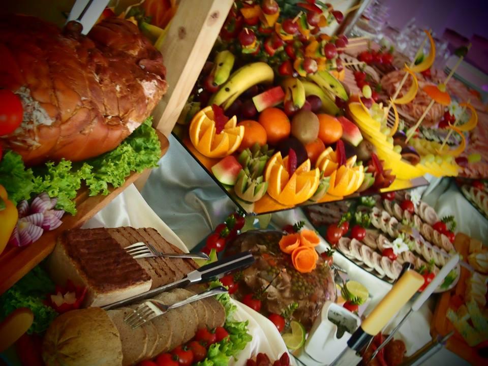 Bogato zastawiony stół z różnorodnymi potrawami: pieczone mięso, wędliny, pieczywo, dekoracje z owoców (banany, pomarańcze, kiwi, winogrona), warzywa i sałatki, tworzące wykwintną kompozycję.