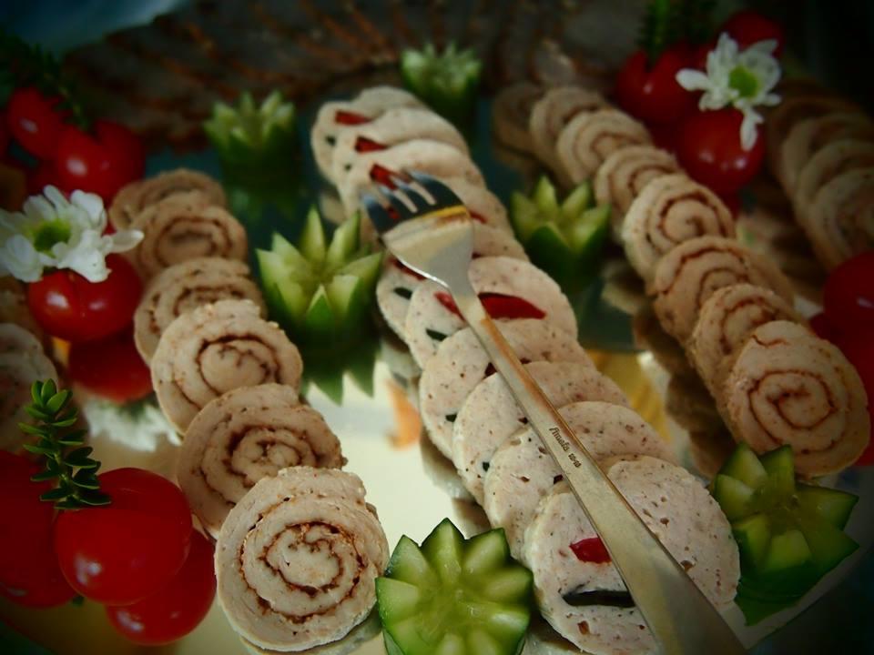 Dekoracyjnie ułożone roladki mięsne i wędlinowe na tacy z pomidorkami koktajlowymi, ogórkami dekoracyjnie wyciętymi w kształcie kwiatów i białymi kwiatami, całość sfotografowana z bliska...