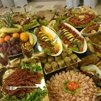 Bogato zastawiony stół z różnorodnymi potrawami: owoce, warzywa, mięsa, sałatki, wszystko starannie ułożone na dużych półmiskach i talerzach. W tle widoczne dekoracyjne elementy sali.