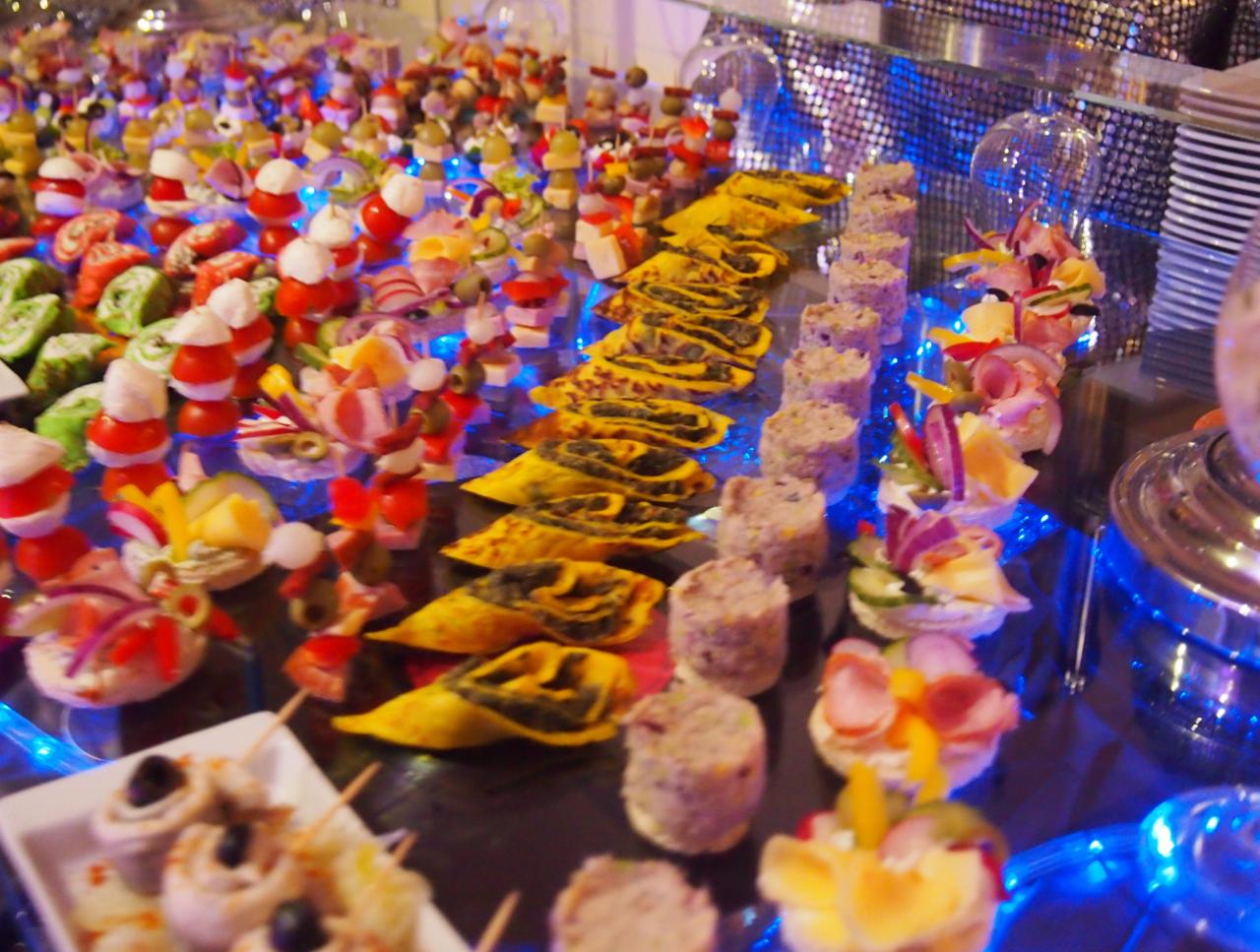 Bogato zastawiony stół z różnorodnymi przekąskami: mini szaszłyki z pomidorków koktajlowych i mozzarelli, roladki z ciasta francuskiego, pasztety w formie walców, tartaletki z wędliną i warzywami...