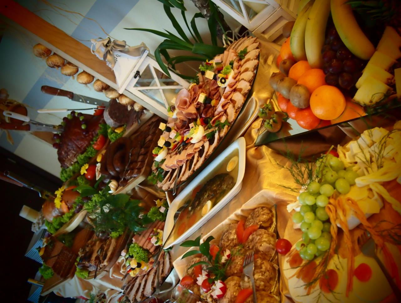 Bogato zastawiony stół z różnorodnymi potrawami mięsnymi, owocami i dekoracjami roślinnymi, widoczne półmiski z wędlinami, pieczonym mięsem, świeże owoce jak banany, mandarynki, winogrona i kiwi...