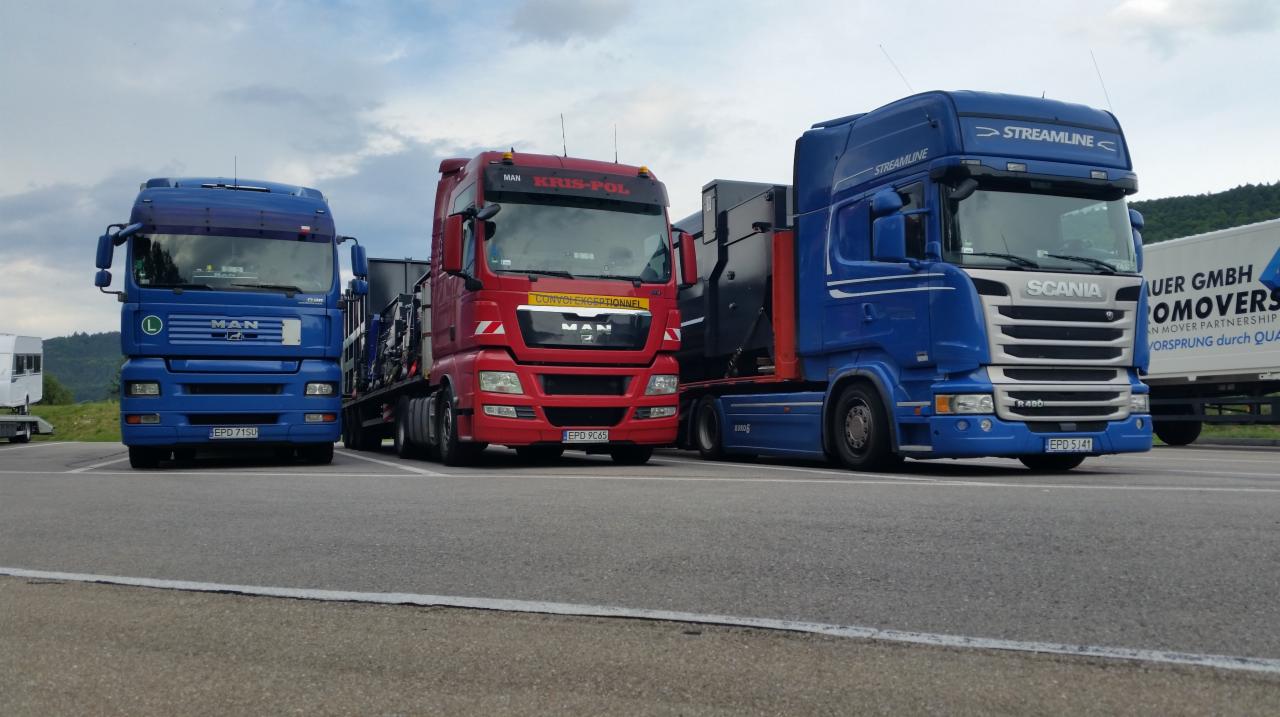 Trzy ciężarówki różnych marek (MAN i Scania) stojące obok siebie na parkingu; pierwsza niebieska, druga czerwona z napisem KRIS-POL, trzecia niebieska z napisem STREAMLINE; w tle widać zalesione...