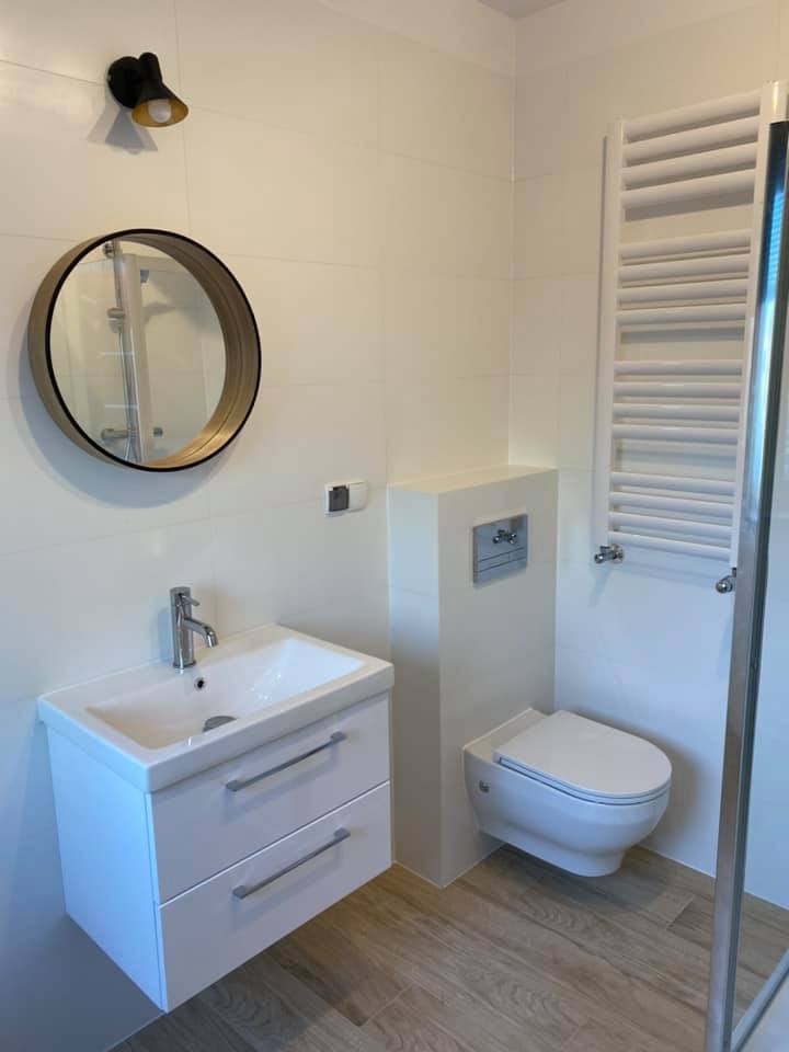 Nowoczesna, jasna łazienka z białymi płytkami, drewnopodobną podłogą, wiszącą szafką z umywalką, okrągłym lustrem w złotej ramie i grzejnikiem drabinkowym.