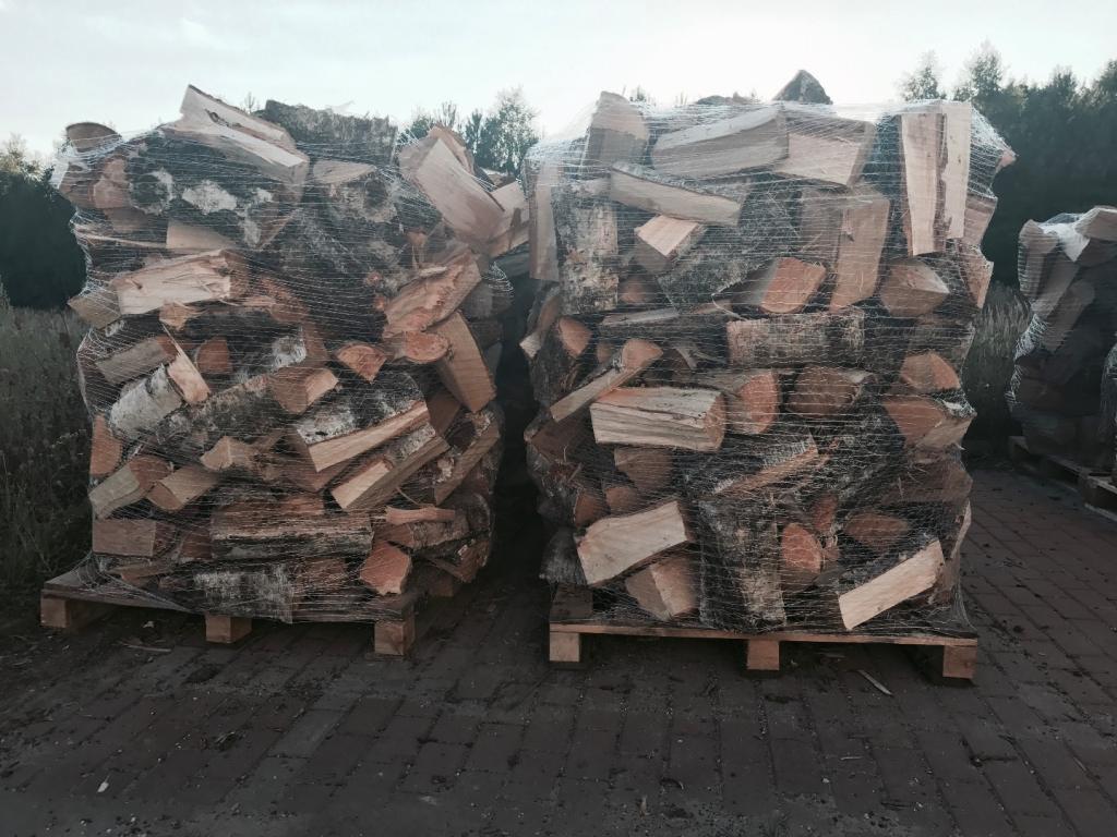 Dwa palety ułożonego, pociętego drewna, owinięte siatką, stoją na zewnątrz na brukowanej powierzchni.