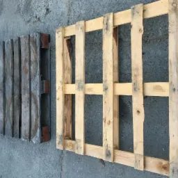 Dwie drewniane palety leżące na betonowej powierzchni: jedna szara, druga z jasnego drewna, ukazujące strukturę drewna i złączenia.