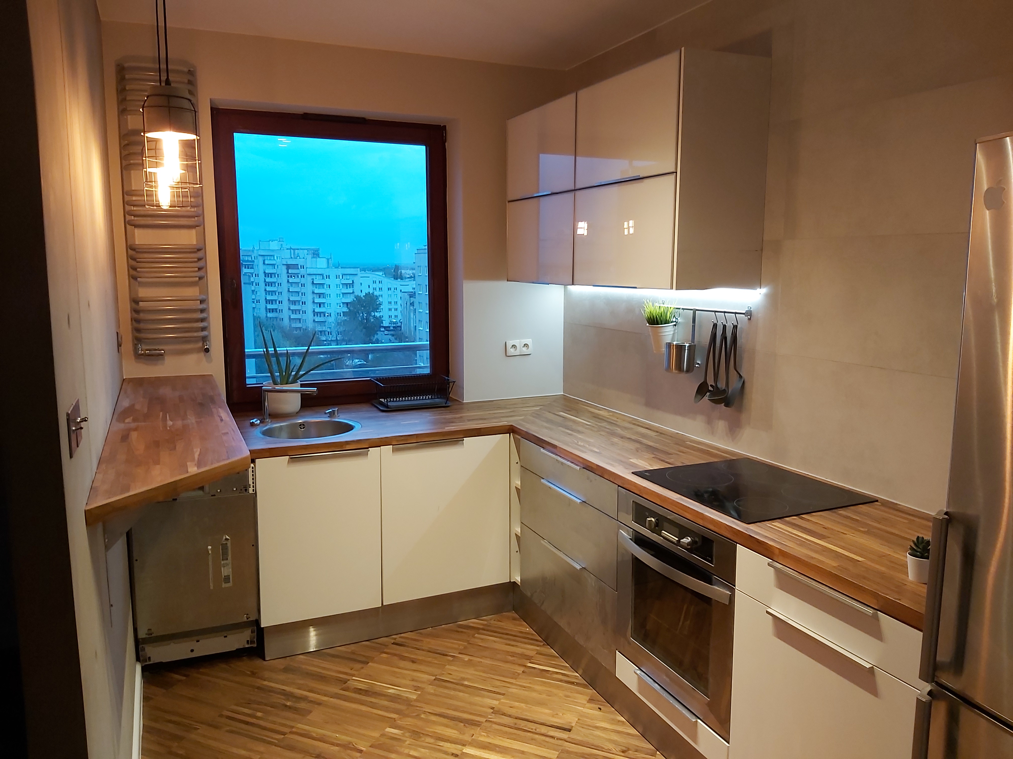 Nowoczesna kuchnia z drewnianym blatem, białymi szafkami i widokiem na miasto za oknem. Nad zlewem widoczny grzejnik drabinkowy, a nad blatem roboczym reling z akcesoriami kuchennymi...