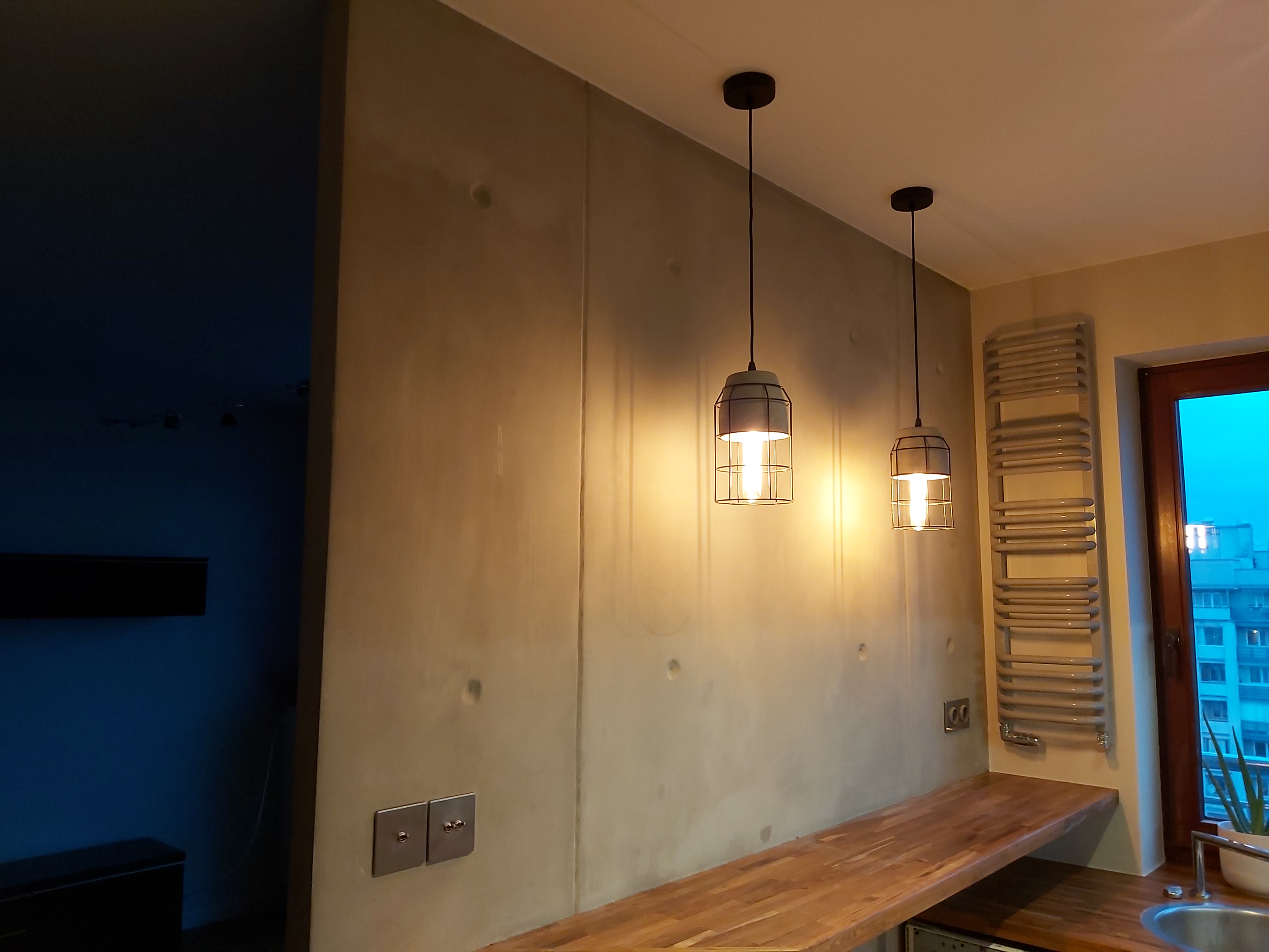 Surowa ściana z betonu architektonicznego w minimalistycznej kuchni, oświetlona dwoma wiszącymi lampami w metalowych kloszach, z drewnianym blatem i grzejnikiem drabinkowym w tle