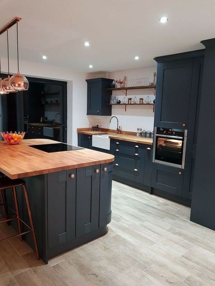 Granatowa kuchnia z drewnianym blatem i wyspą, miedziane lampy wiszące, jasna podłoga imitująca drewno, styl klasyczny z nowoczesnymi akcentami.