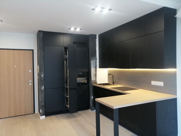 Nowoczesna kuchnia w czerni z drewnianym blatem, oświetleniem LED podszafkowym i otwartą szafką cargo, widoczna na tle jasnej podłogi i drzwi wejściowych.