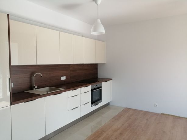 Nowoczesna kuchnia z białymi lakierowanymi szafkami, drewnianym blatem i brązowym panelem ściennym, widok na zlewozmywak i piekarnik, podłoga wyłożona panelami imitującymi drewno.