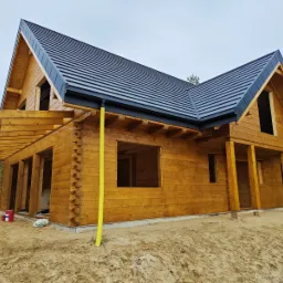 Surowy, dwukondygnacyjny dom z bali z czarnym dachem i żółtą rurą spustową, w trakcie budowy, widoczne niezabudowane otwory okienne i drzwiowe, teren wokół domu piaszczysty, w tle las.