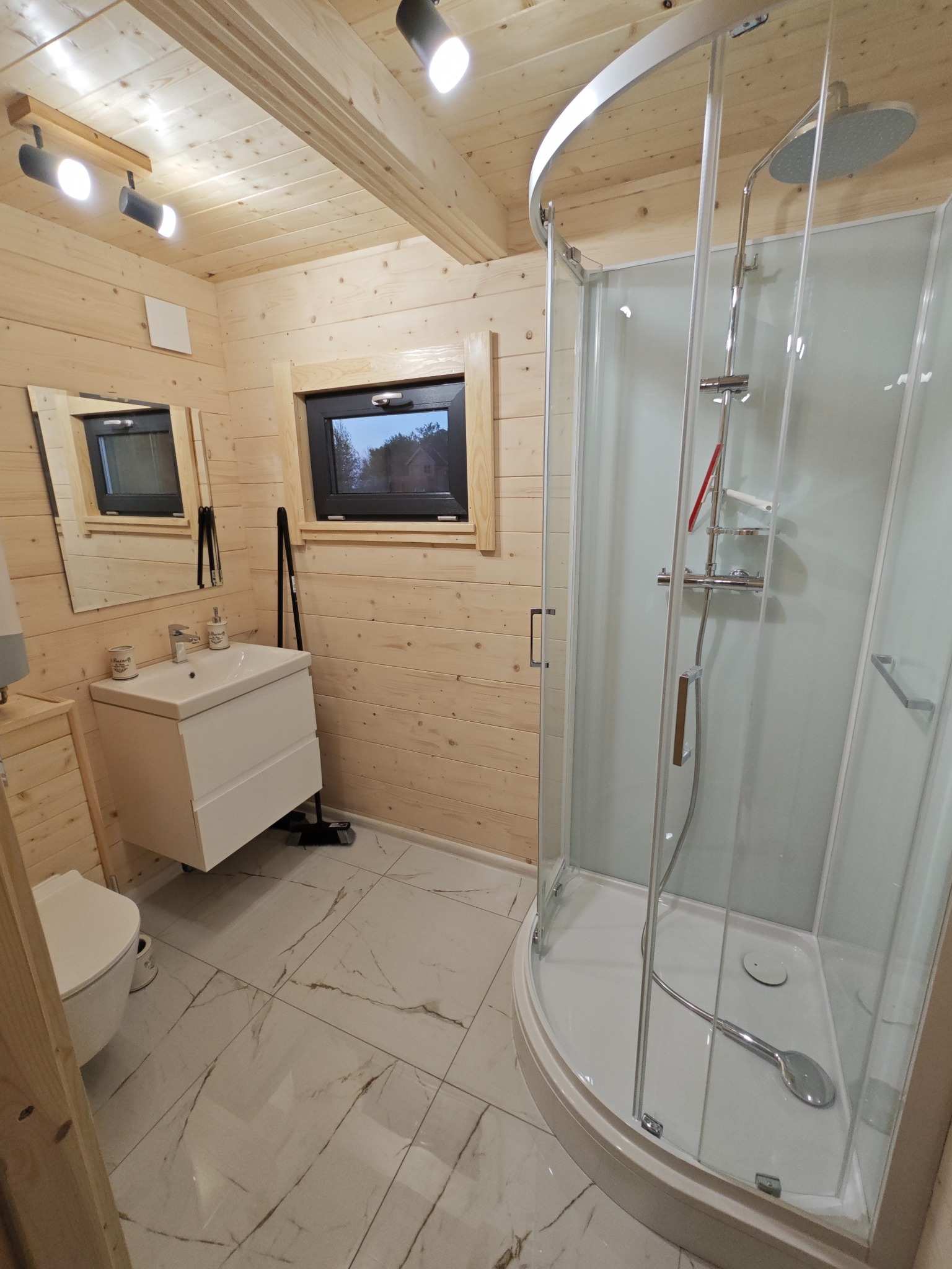 Wnętrze łazienki w domu z bali: kabina prysznicowa z hydromasażem, biała umywalka z szafką, lustro i toaleta na marmurowej podłodze, oświetlenie punktowe.