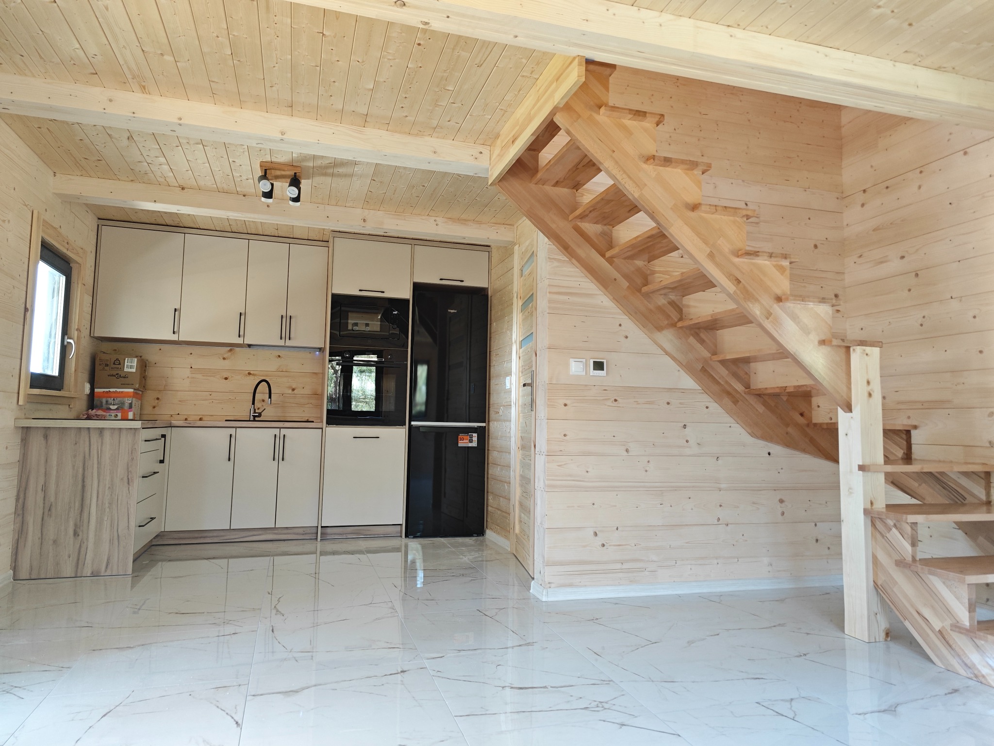 Wnętrze domu z bali: jasna kuchnia z nowoczesną zabudową, drewniane schody na piętro, białe płytki podłogowe z marmurkowym wzorem, ściany i sufit wykończone jasnym drewnem.