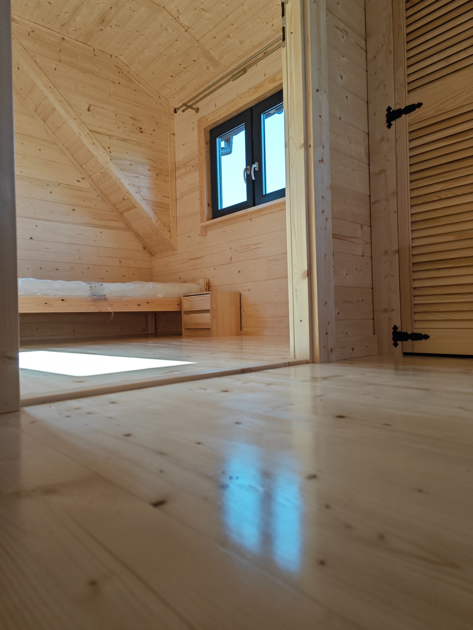 Wnętrze drewnianego domu z bali, widok z niskiej perspektywy na błyszczącą podłogę, w tle łóżko z materacem w folii i szafka nocna, czarne okno w ramie i drzwi z żaluzjami z ozdobnymi zawiasami.