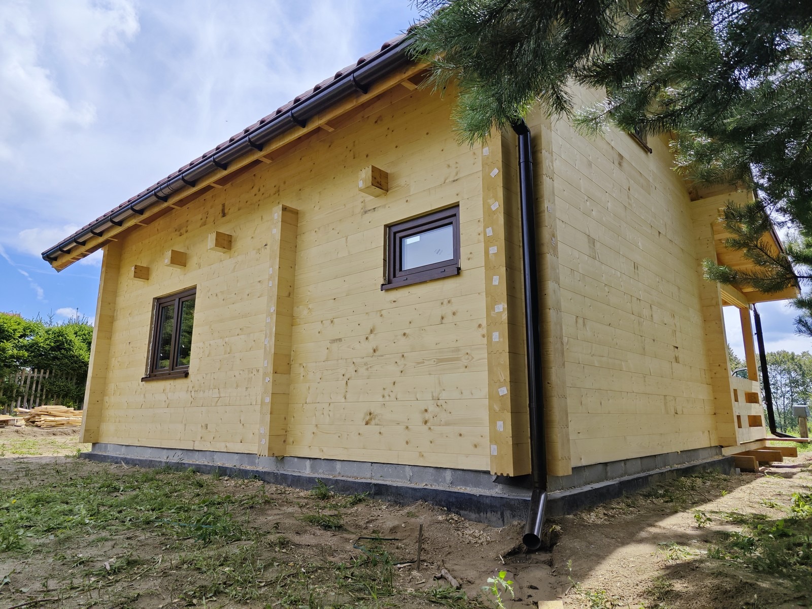 Surowy stan drewnianego domu z bali z ciemnymi ramami okiennymi i rynnami, widoczne tymczasowe zabezpieczenia na łączeniach belek, częściowo zadaszony taras z boku.