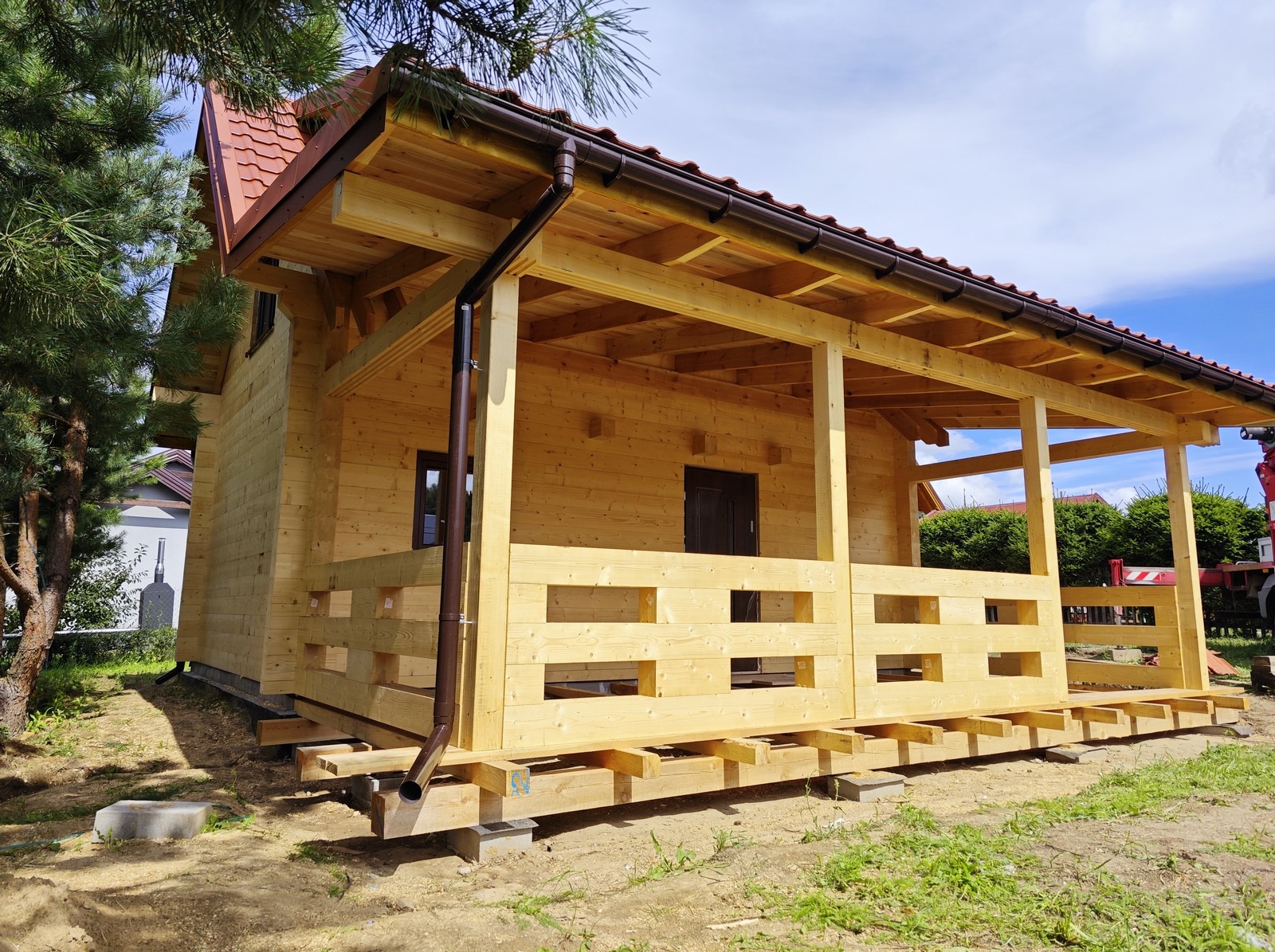 Surowy drewniany dom w trakcie budowy, widoczny taras z balustradą, brązowe rynny, dom posadowiony na betonowych bloczkach, w tle błękitne niebo z chmurami i zieleń drzew.