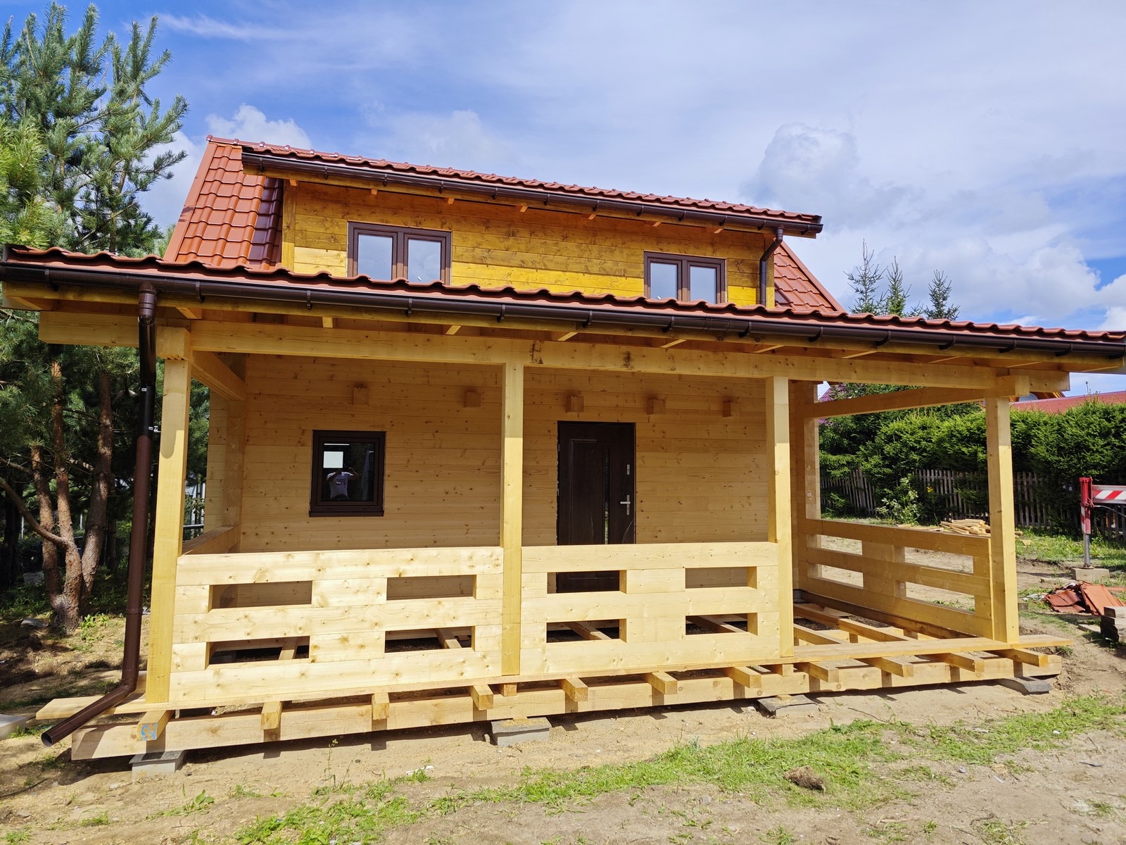 Surowa konstrukcja drewniana z brązowym dachem, widoczne elementy tarasu i przygotowane podłoże, w tle zieleń drzew i krzewów.