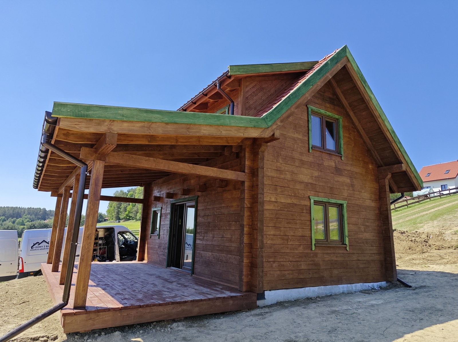 Gotowy dom z bali z zadaszonym tarasem, pomalowany na brązowo z zielonymi akcentami, na tle błękitnego nieba i zieleni.