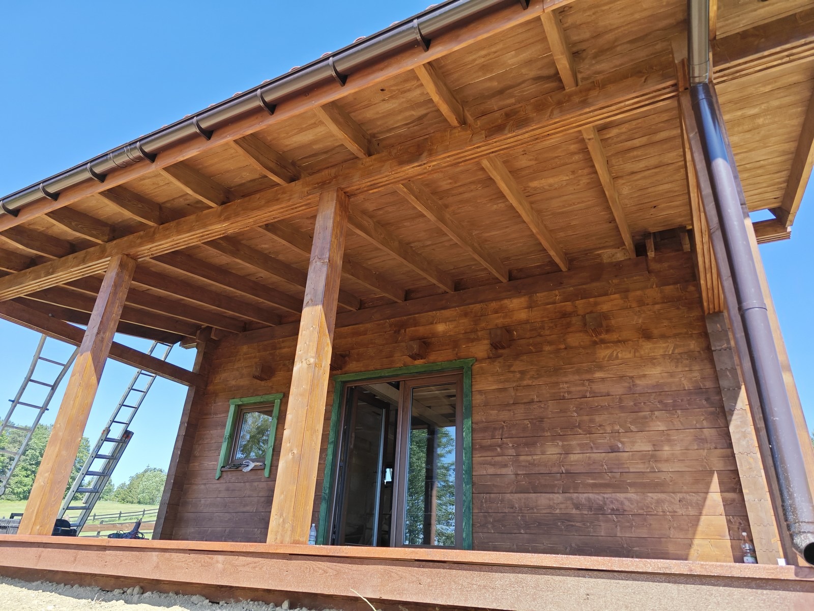Surowa drewniana konstrukcja domu z bali z widocznym tarasem, zielonymi ramami okiennymi i drabiną opartą o ścianę boczną.