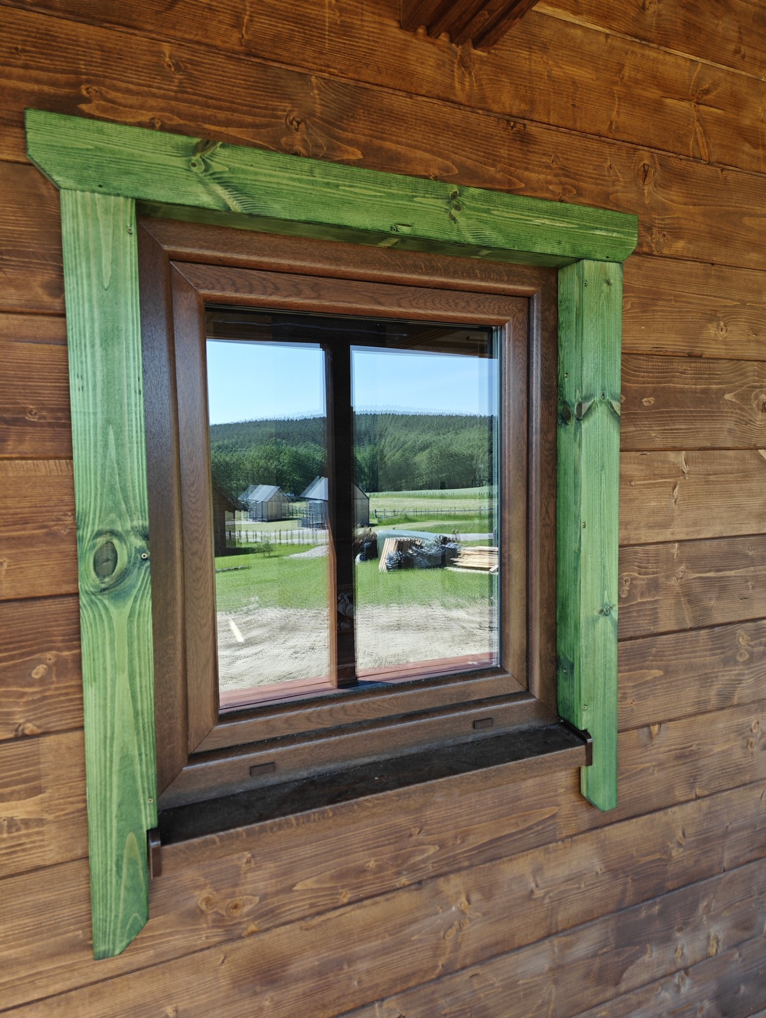 Okno z brązową ramą i zieloną, drewnianą dekoracją na ścianie z brązowych bali. W szybie okna odbija się krajobraz z zabudowaniami i drzewami.