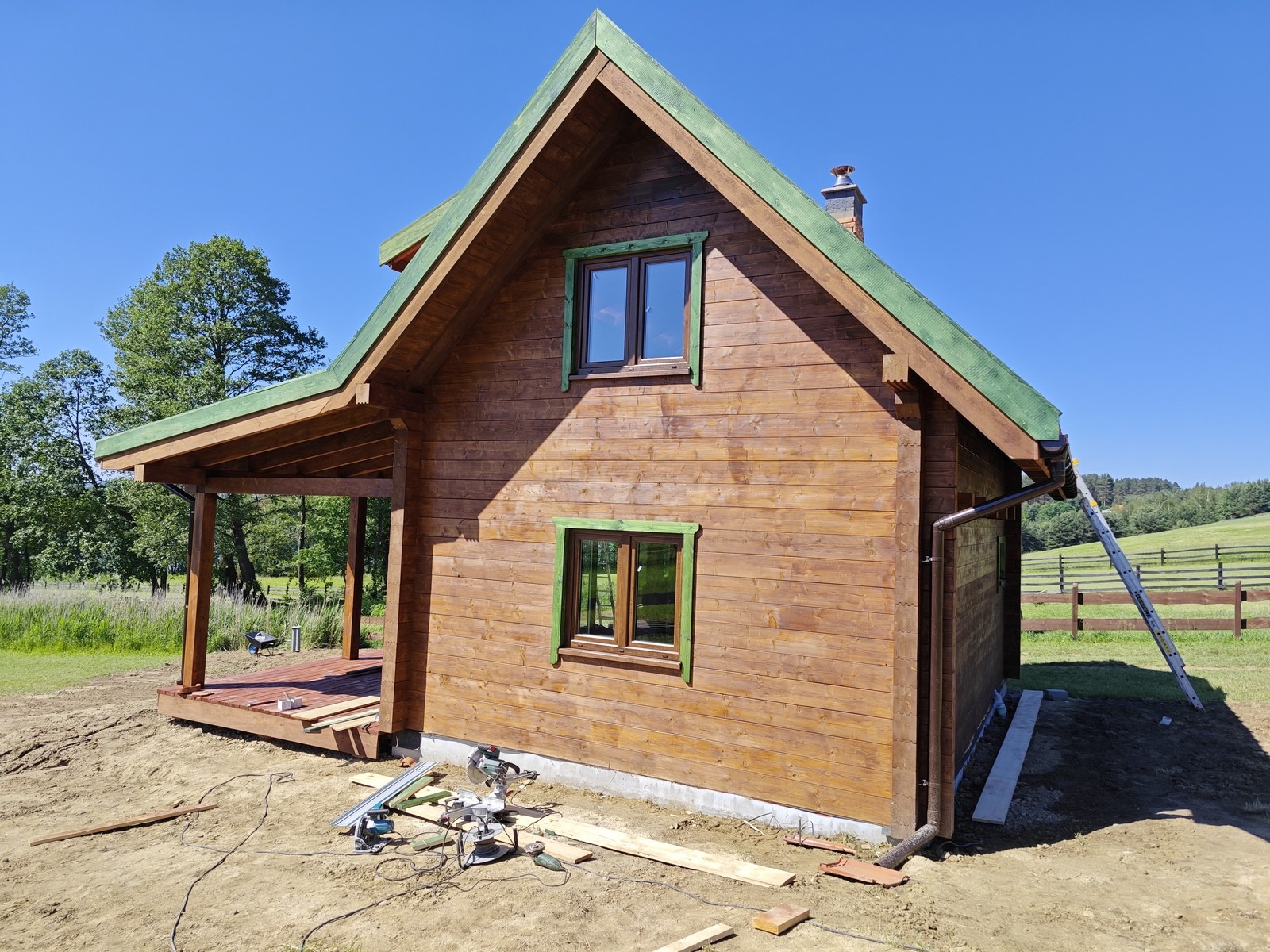 Brązowy dom z drewna z zielonymi akcentami na dachu i oknach, widoczne narzędzia budowlane i drabina oparta o rynnę, w tle zielone łąki i błękitne niebo.