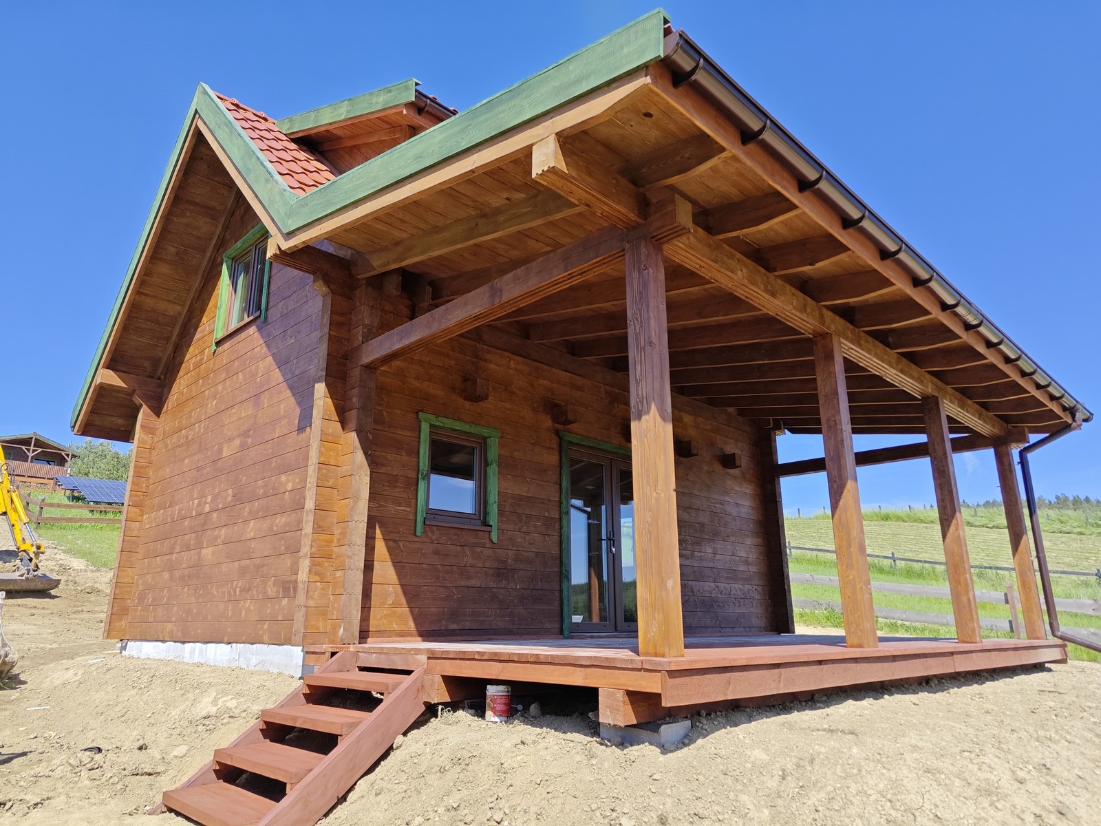 Brunatny drewniany dom z bali z zielonymi elementami wykończeniowymi, tarasem i schodami wejściowymi, częściowo wykończony teren wokół, błękitne niebo w tle.