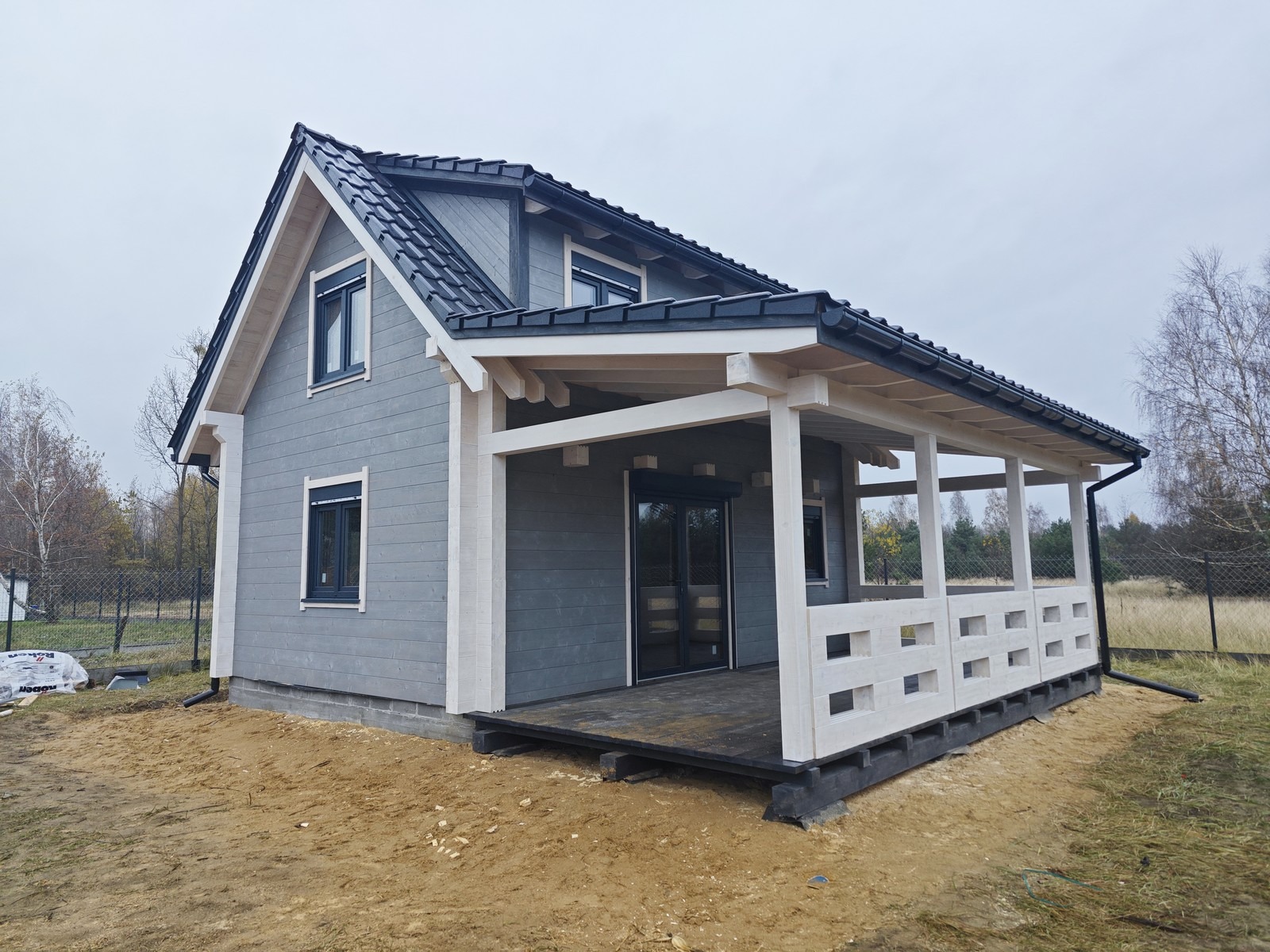 Szary dom z bali z białymi detalami i zadaszonym tarasem, widok z zewnątrz na tle jesiennego krajobrazu, widoczne worki z materiałami budowlanymi obok budynku.