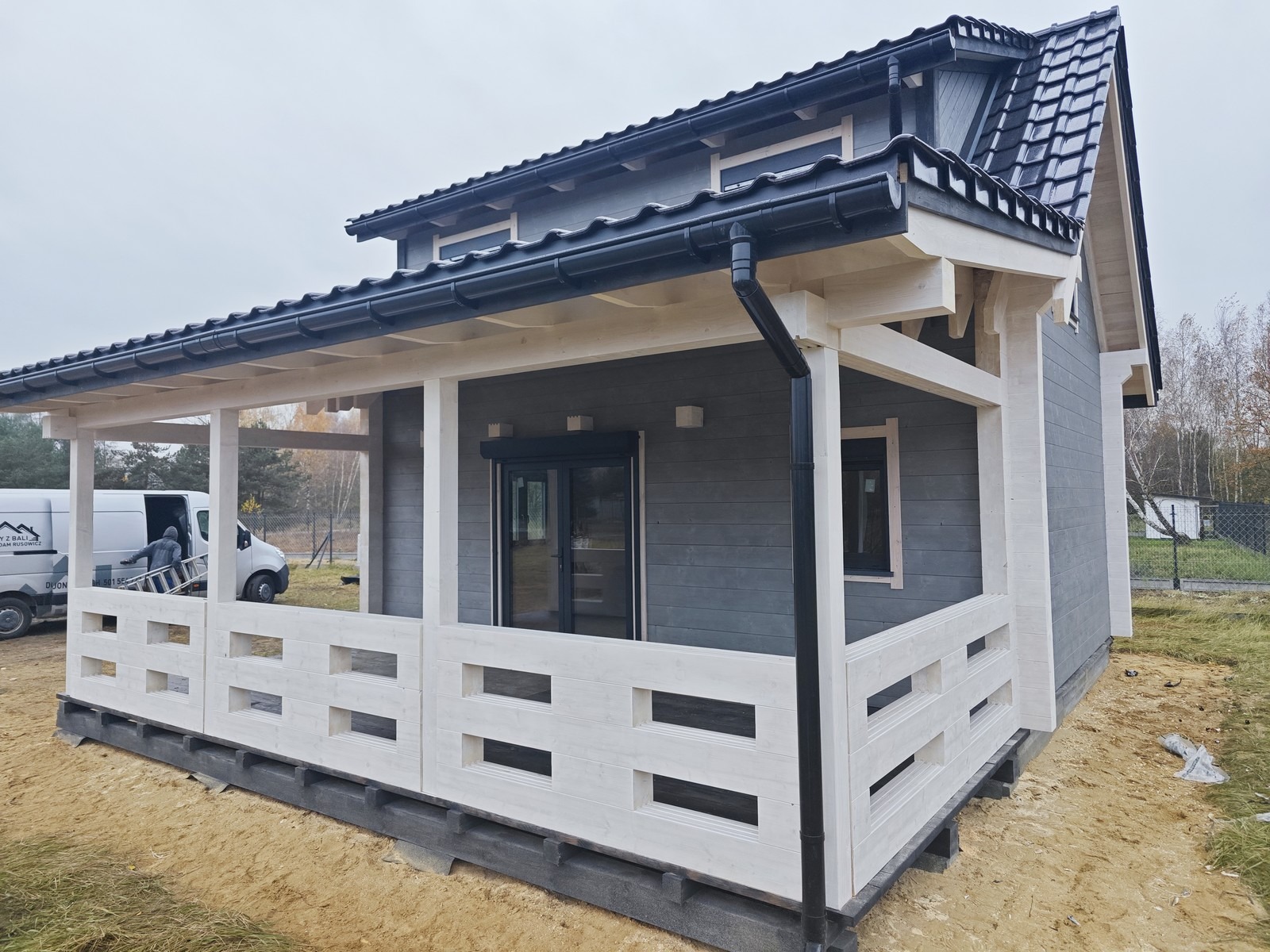 Nowoczesny dom z bali z tarasem w Mrągowie, szara elewacja, białe elementy konstrukcyjne, widoczny samochód dostawczy firmy w tle.