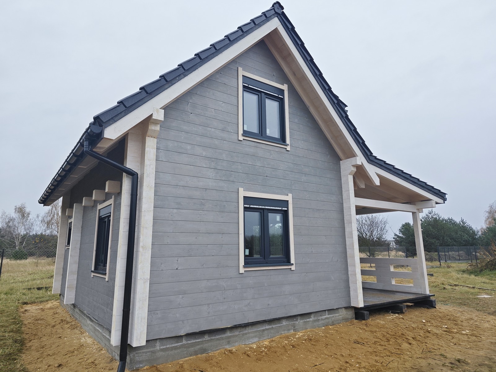Szary, drewniany dom z bali z czarną dachówką i białymi detalami wykończeniowymi, widoczny z boku, z małym zadaszonym tarasem z przodu. Dwa okna z czarnymi ramami.
